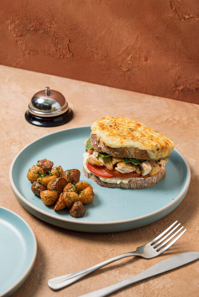 Sandwich de pan artesanal con pollo, tomate y queso gratinado dorado, acompañado de papas rústicas en un plato celeste.