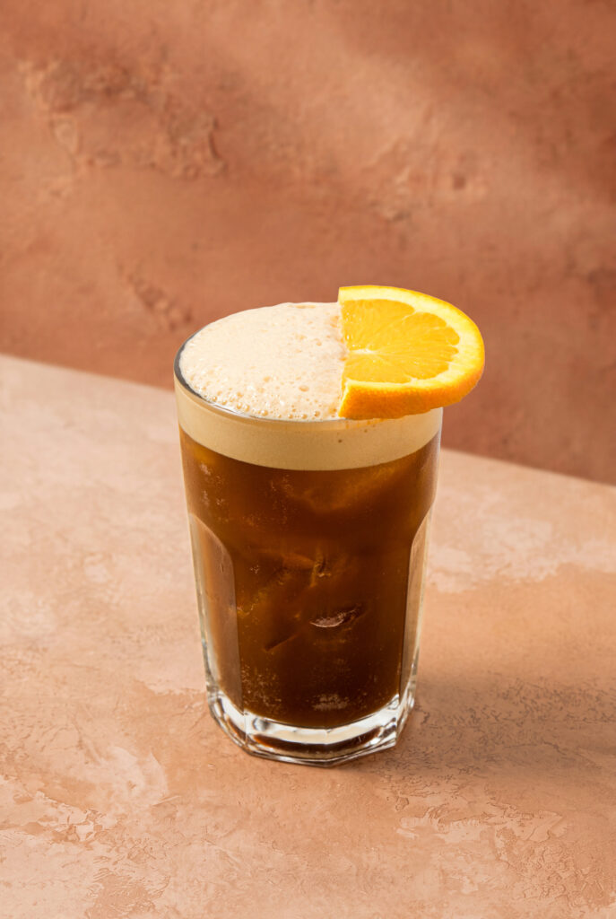 Vaso de cóctel carajillo con espuma cremosa en la parte superior y una rodaja de naranja como decoración.