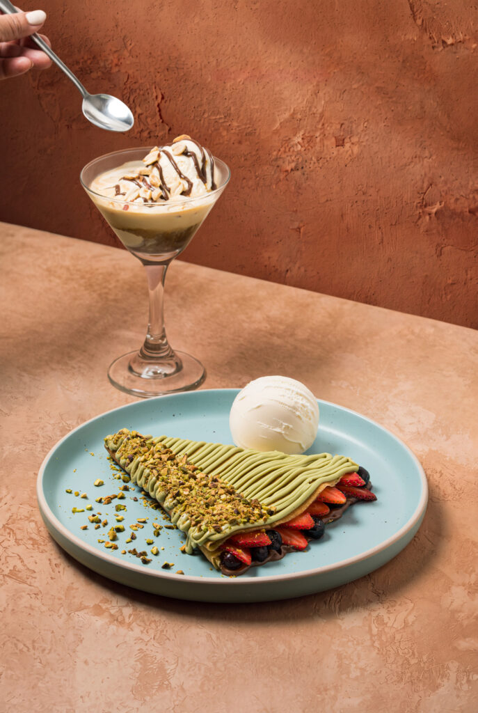 Mano sirviendo con cuchara un affogato martini junto a una crepa de pistacho con frutos rojos.