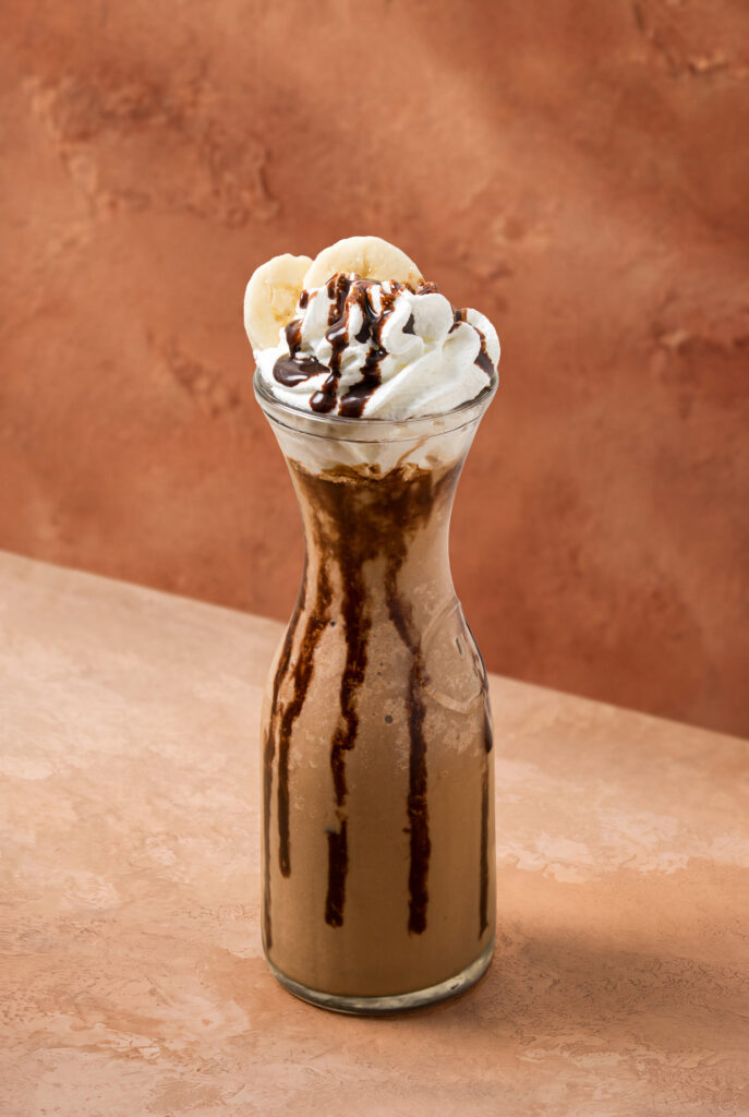 Jarra de vidrio con frappé de Nutella y banano, coronado con crema batida, rodajas de banano y salsa de chocolate escurriendo por los lados.
