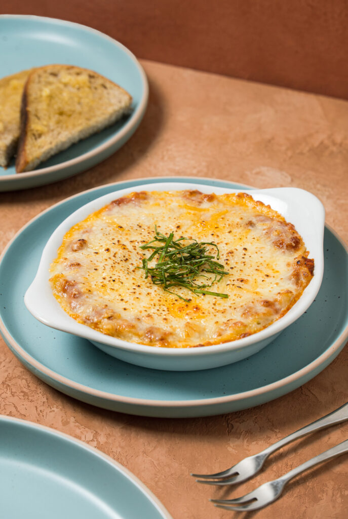 Cazuela individual con lasaña a la boloñesa cubierta de queso gratinado dorado, acompañada de pan tostado.