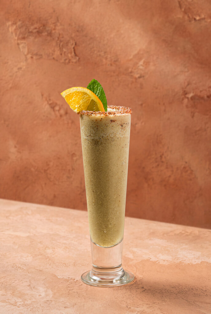 Vaso alto de naranjada chapina con borde escarchado de especias, decorado con rodaja de naranja y hoja de menta.